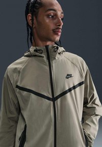 Beige lättviktsjacka med huva, med svarta accenter och ett diagonalt randmönster. Dragkedja fram och sidofickor. Nike-logotypen är närvarande.