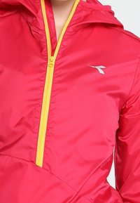 Red windbreaker with a yellow zipper and hood. Features a silver logo on the chest. Material appears lightweight and slightly shiny.