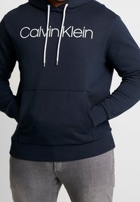 Felpa con cappuccio blu navy con stringhe bianche e logo "Calvin Klein". Presenta una tasca frontale e polsini e orlo a coste.