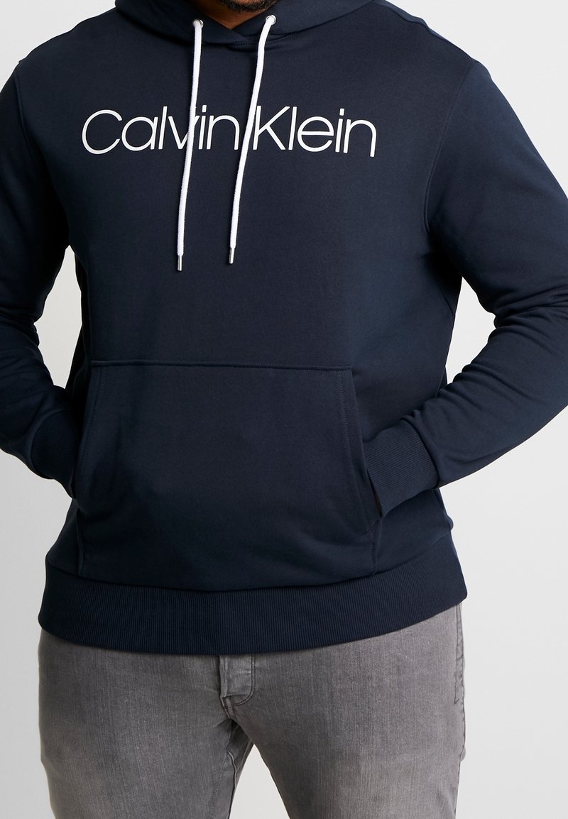Felpa con cappuccio blu navy con stringhe bianche e logo "Calvin Klein". Presenta una tasca frontale e polsini e orlo a coste.