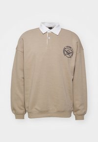 Beżowy sweatshirt z białym kołnierzykiem, długimi rękawami i nadrukowanym logo na lewym czole, o luźnym kroju i miękkiej fakturze.