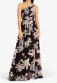 Femme portant une robe maxi noire à une épaule avec un grand motif floral blanc et beige, debout les bras le long du corps.