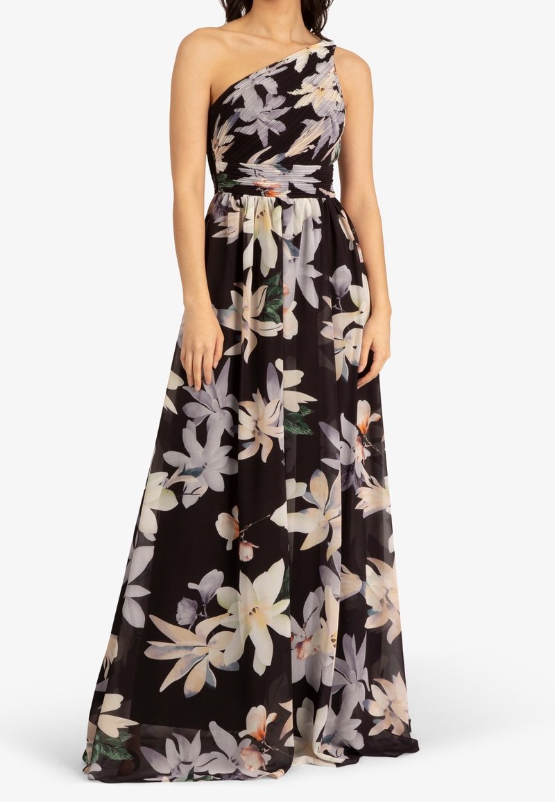 Femme portant une robe maxi noire à une épaule avec un grand motif floral blanc et beige, debout les bras le long du corps.