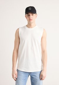 Camiseta sin mangas de algodón blanca con cuello redondo, combinada con jeans azul claro. Se lleva una gorra negra con acentos de texto en blanco.