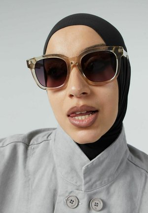 Oversized zonnebril met een heldere, lichtbruine montuur en donkere lenzen. Textuurafwerking met brede glazen. Model draagt een zwarte hijab.