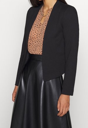 Femme portant un blazer noir sur un chemisier marron à pois et une jupe en cuir noire, accessoirisée d’un collier chaîne en or.