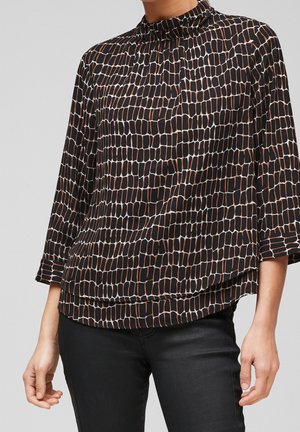 Femme portant un chemisier noir ample avec un motif rectangulaire abstrait marron et blanc et un pantalon noir sur fond uni.