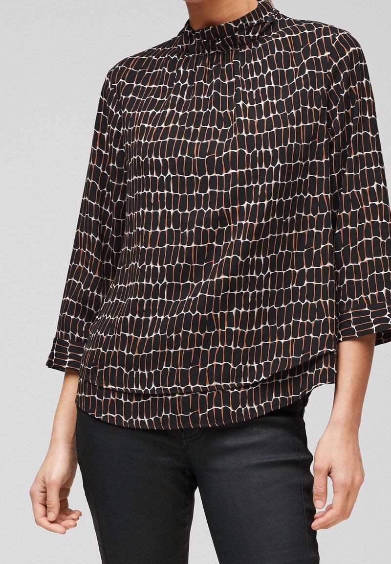 Femme portant un chemisier noir ample avec un motif rectangulaire abstrait marron et blanc et un pantalon noir sur fond uni.