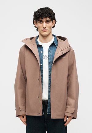 Ung mand med krøllet hår iført en beige hættetrøje over en denimskjorte og en hvid T-shirt med grafik, stående foran en ensfarvet baggrund.