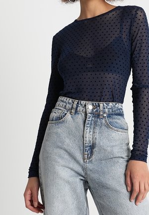 Vrouw draagt een marineblauw transparant polkadot shirt met lange mouwen en een bijpassende beha eronder, gecombineerd met lichtblauwe high-waisted spijkerbroek.