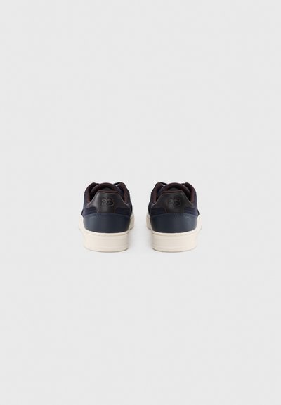 Ζευγάρι από ναυτικό μπλε αθλητικά παπούτσια με λευκές σόλες, απεικονιζόμενα από πίσω, με το λογότυπο "PS" στην ταμπέλα της φτέρνας, πάνω σε λευκό φόντο.