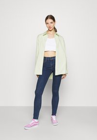 Camisa oversized verde clara, top cropped branco, jeans skinnies azul escuro e ténis cor-de-rosa com cordões brancos. Texturas suaves.