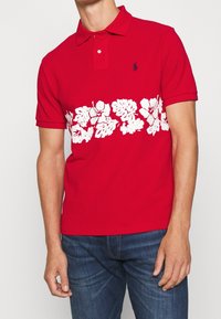 Polo rouge avec un motif floral blanc sur la poitrine, manches courtes et col classique. Fabriqué en coton lisse.