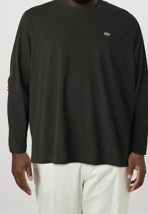 Personne portant un haut à manches longues noir avec un petit logo crocodile vert sur la poitrine et un pantalon blanc, debout devant un fond uni.