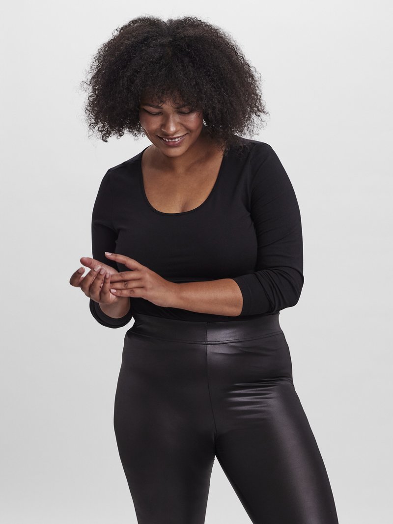 Μαύρο πουκάμισο με μακριά μανίκια και στρογγυλή λαιμόκοψη, σε συνδυασμό με γυαλιστερά μαύρα κολάν. Το μοντέλο έχει σγουρά μαλλιά και κοιτάζει τα χέρια της.