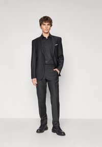 Un costume noir avec une veste sur mesure et un pantalon assorti, complété par une chemise sombre et des chaussures noires, présentant un design élégant et une texture lisse.