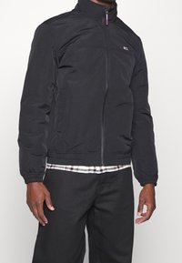 Man som bär en svart zip-up jacka med elastiska ärmar och en liten logotyp på bröstet, ihop med svarta byxor och en rutig skjorta som sticker fram.