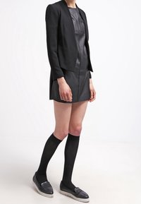 Černý strukturovaný blejzr přes krátké kožené šaty, doplněný o černé podkolenky a texturované loafers, s elegantním designem a moderním střihem.
