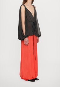 Blouse plissée noire avec un décolleté en V profond et des manches amples, associée à une jupe plissée orange vif, rehaussée d'un nœud noir à la taille.