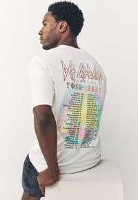 White cotton t-shirt featuring colorful "Def Leppard Tour 1983" graphic with city names. Short sleeves and relaxed fit with soft texture.