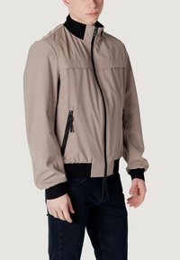 Giacca beige chiaro con colletto alto, chiusura a zip frontale e due tasche con zip. Presenta dettagli neri sul colletto e sui polsini.