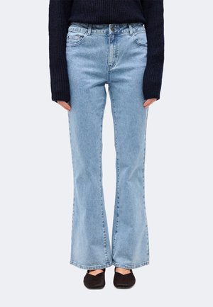 Personne portant un jean flare taille haute bleu clair, un pull en maille noir avec des manches couvrant les mains, et des chaussures plates noires, debout devant un fond uni.