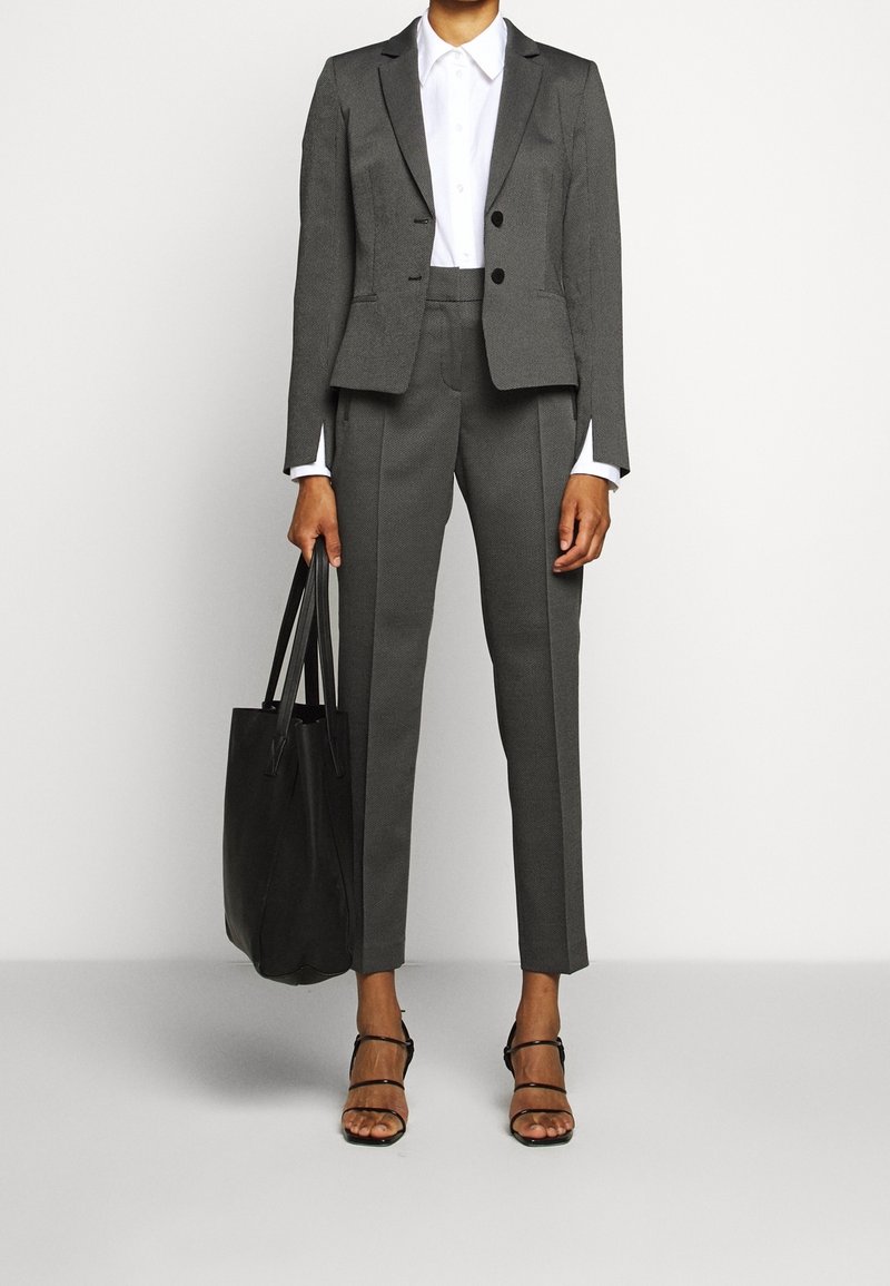 Femme portant un tailleur gris foncé sur mesure avec pantalon court, chemise blanche boutonnée, talons noirs à brides, et tenant un grand sac cabas noir.