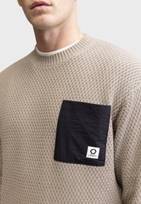 Maglione lavorato a maglia beige con un motivo testurizzato, colletto a costine e una tasca a toppa nera con etichetta logo sul lato sinistro in alto.