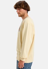 Light yellow sweatshirt with ribbed cuffs and hem. Features an embroidered graphic on the chest. Soft texture and relaxed fit.