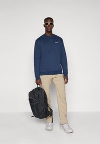 Man met een zonnebril, marineblauwe sweatshirt, beige broek en witte sneakers, met een zwarte rugzak tegen een effen achtergrond.