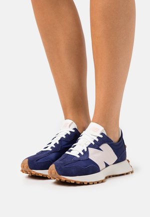 Legs wearing blue and white New Balance sneakers with gum-colored soles, standing on a white surface.