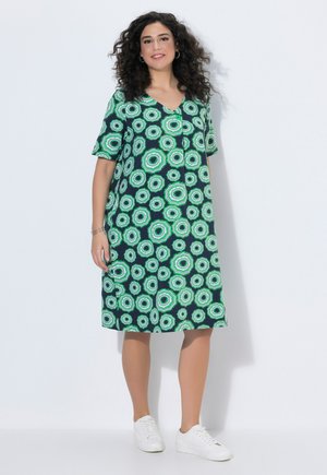 Frau mit lockigem Haar, die ein knieumspieltes, grün und dunkelblau gemustertes Kleid und weiße Turnschuhe trägt, vor weißem Hintergrund stehend.