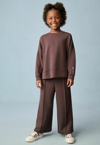 Brązowy sweter z opadającymi ramionami i haftowanym detalem, w połączeniu z szerokimi brązowymi spodniami. Białe sneakersy z wzorem dopełniają całość.