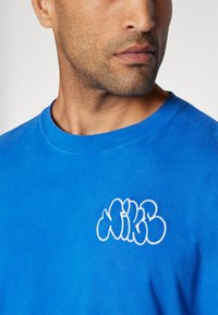 Camiseta de algodón azul con un prominente logo blanco bordado en el pecho. Cuello redondo y mangas cortas con una textura suave.