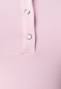 Lys pink, tætsiddende beklædningsgenstand med en tekstureret overflade og tre metal snap-knapper på fronten, der skaber et enkelt, moderne design.