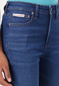 Jeans de mezclilla azul oscuro con un bolsillo frontal que presenta un logo de Calvin Klein en rojo y blanco, costuras resistentes y un diseño clásico de cinco bolsillos.