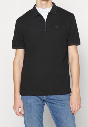 Homme portant un polo noir à manches courtes avec une fermeture éclair et un jean bleu clair sur un fond uni.