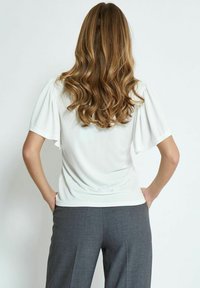 Top bianco con maniche corte a sbuffo, realizzato in materiale morbido. Abbinato a pantaloni grigi su misura. Il retro del top è liscio e aderente.