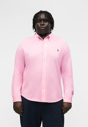 Vyras su dredais, dėvintis šviesiai rožinę ilgomis rankovėmis marškinių su saga ir tamsias kelnes, stovintis priešais vienspalvį foną.