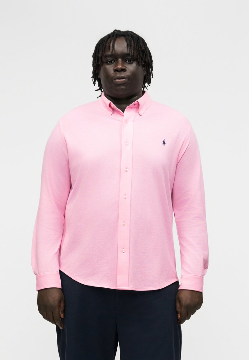 Mann med dreadlocks iført en lys rosa langermet skjorte med knapper og mørke bukser, stående mot en ensfarget bakgrunn.