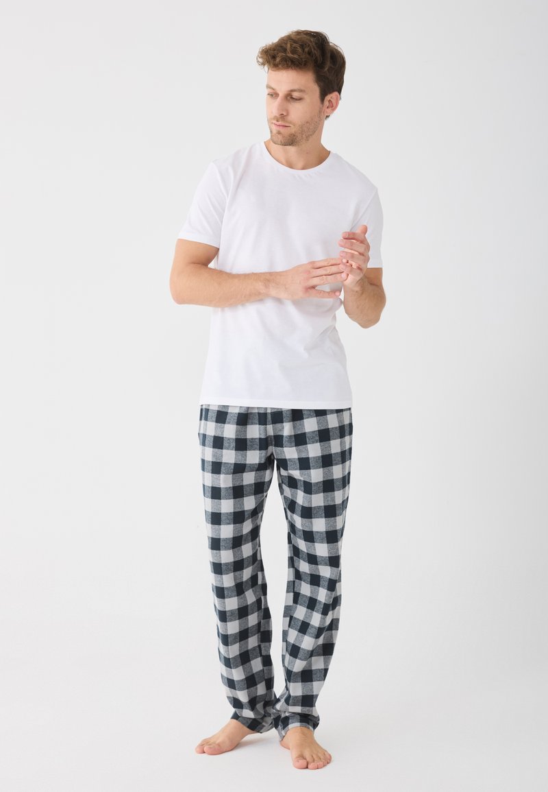 Biela krátkorpíšová tričko v kombinácii s čierno-bielymi károvanými pyžamovými nohavicami, so voľným strihom a mäkkou textúrou. Postoj naboso.