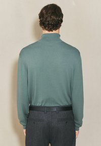 Pulover tricotat verde cu nervuri, cu guler înalt, asortat cu pantaloni gri cu dungi și o curea neagră. Textură netedă și croială lejeră.