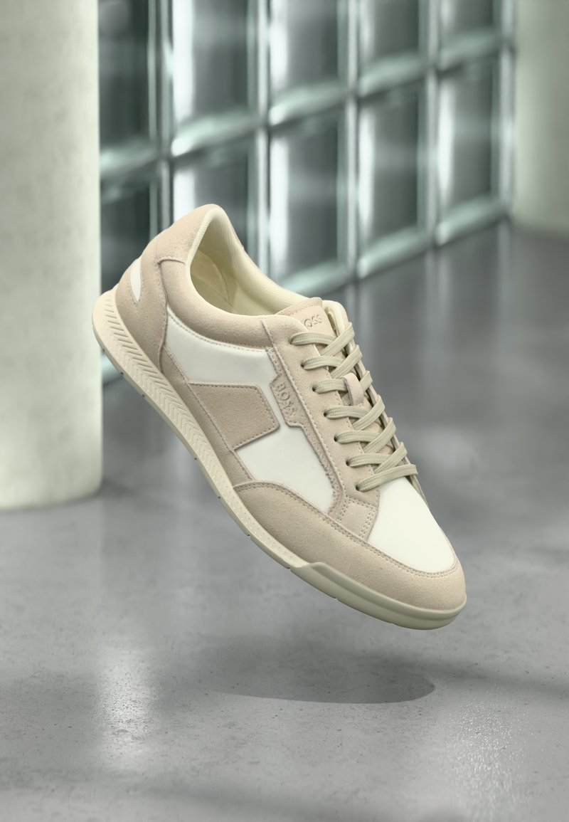 Baskets décontractées beige et blanches avec lacets, panneaux en daim et en cuir, flottant au-dessus d'un sol en béton poli près de blocs de verre.