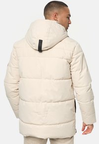 Beige pufferjakke med hætte, der har quiltet struktur, elastiske manchetter og en sort detalje bagpå. Holdbart materiale til varme.