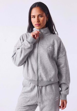 Mujer con chaqueta y pantalones deportivos a juego en gris claro con cremallera y logo, sosteniendo la cremallera de la chaqueta cerca de su barbilla.