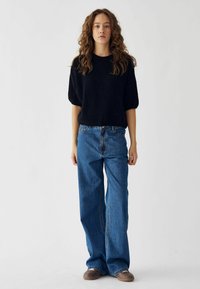 Čierny pletený sveter s krátkymi, širokými rukávmi; vysoko pásové, voľné modré džínsy; hnedé topánky; jednoduché pozadie.