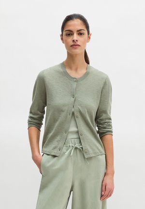 Frau mit zurückgebundenem dunklem Haar, die eine hellgrüne, geknöpfte Strickjacke über einem passenden, gerippten Tanktop und Kordelhose trägt, steht mit einer Hand in der Tasche.