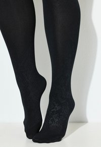 Gambe in collant opachi neri con motivo testurizzato, in piedi sulle punte contro uno sfondo bianco semplice.