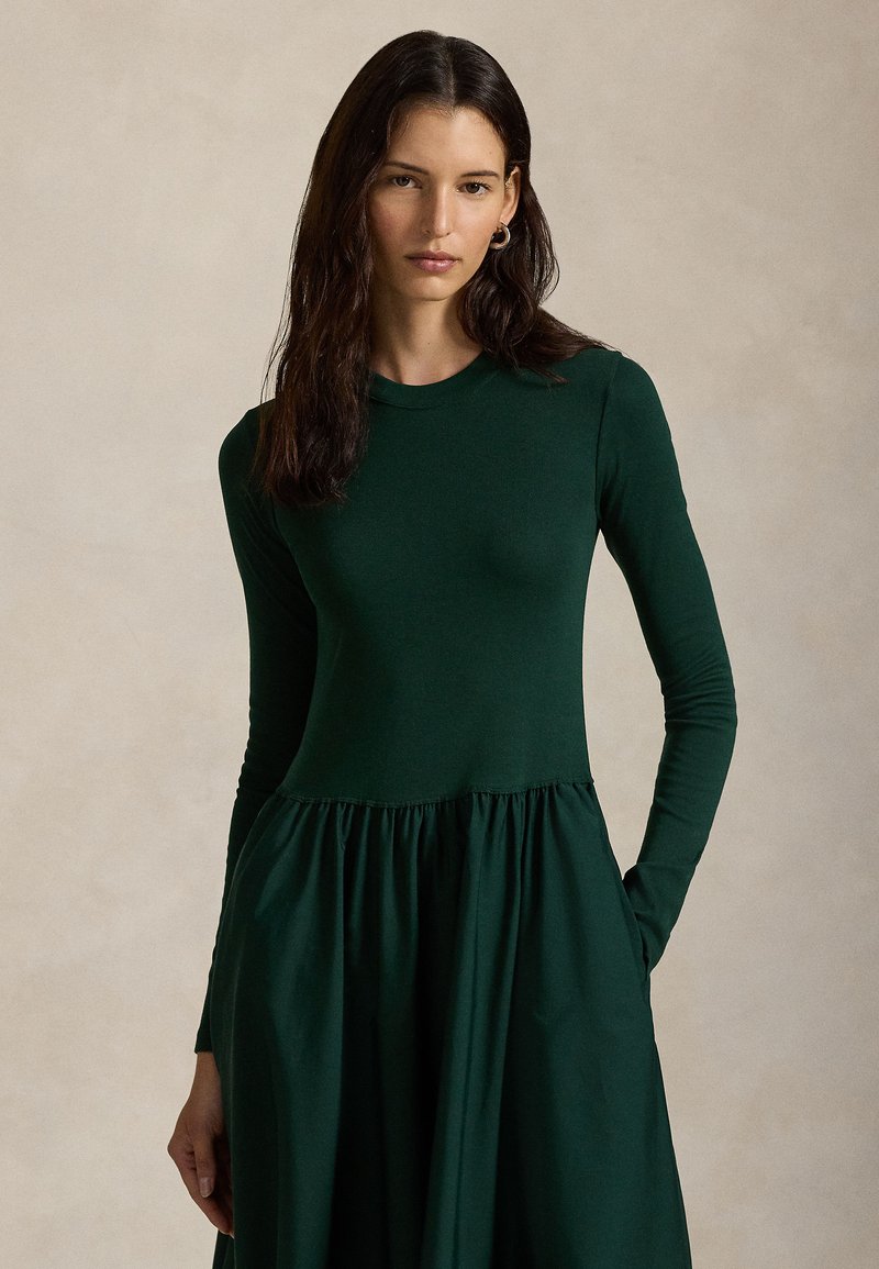 Långärmad, mörkgrön klänning med en figurnära överdel och en flödande kjol med rynkade detaljer vid midjan. Mjuk tygstruktur.