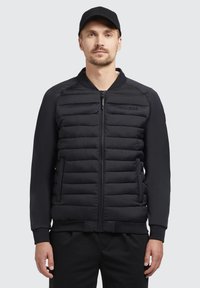 Giacca a pena nera con zip frontale, dotata di sezioni imbottite, polsini a coste e un tessuto liscio sulle maniche. Logo del brand impresso sul petto.